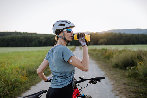 Diabetic cyclist with a continuous glucose monitor on her arm drinking water during her bike tour to manage her diabetes while exercising. Concept of exercise and diabetes.