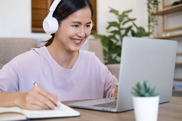 istock Happy young woman learn language listen lecture watch webinar write notes look at laptop sit in living room , Girl student wear wireless headphone study online with skype teacher, distance education, 1697012368