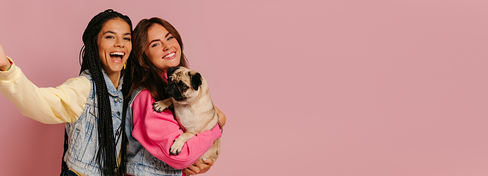 Two joyful young women carrying cute pug dog and making selfie against pink background