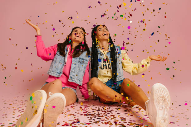 istock Two joyful young women throwing colorful confetti while sitting against pink background 1697014105