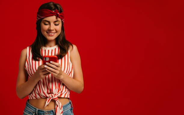 istock Stylish young hipster woman texting message on smart phone and smiling while standing on red background 1697042289