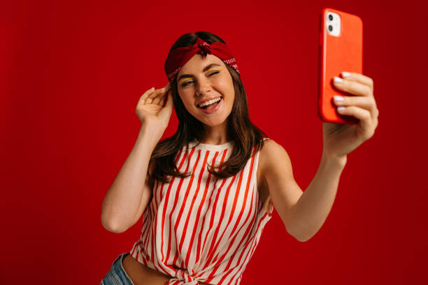 istock Happy young hipster woman making selfie by mart phone and winking while standing on red background 1697042868