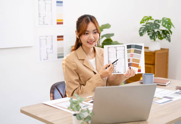 istock Interior designer talking with client on video call, showing architecture blueprint of residence and pantone. Asian architect woman sitting in office, looking at laptop display with online conference. 1697060387