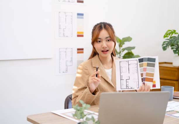 istock Asian Interior designer sitting in front of laptop talking with client on video call, showing architecture blueprint of residence and pantone. business and modern tech, meeting and online conference. 1697060947