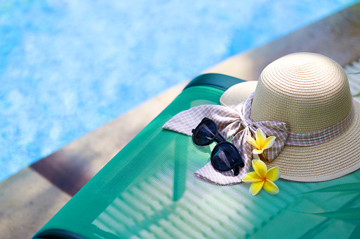 A hat, sunglasses, and a flower petal are resting on a deck chair by the swimming pool, basking in the warm sunshine. This image represents the essence of summer time fun, essential items for a vacation, and the concept of tourism. It embodies the idea of getting away from it all and enjoying a well-deserved holiday.