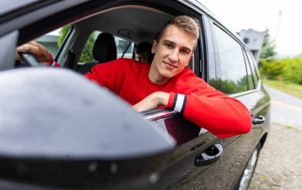 Smiling Teenage Boy Leaning Out of Car A teenage driver, wearing a red sweatshirt, leaning out of a car window and making eye contact with the camera. teenager boy driving stock pictures, royalty-free photos & images
