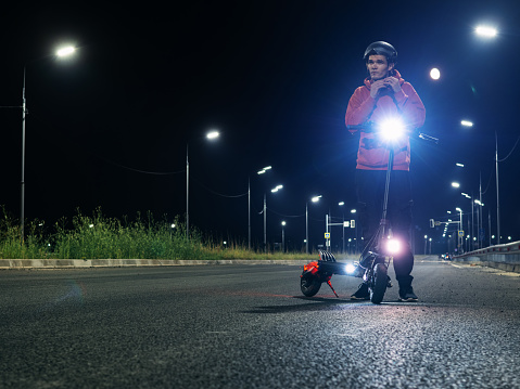 a man puts on a helmet to ride an electric scooter. Night road illuminated by lanterns