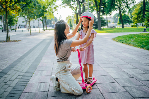 Young mother is adjusting the helmet on her daughter's head. She is smiling and getting ready to ride a scooter in the park
