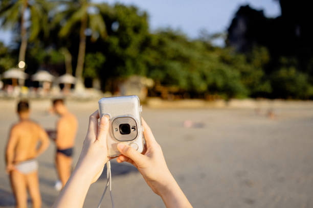 Side wide view Young influencer Asian Using smartphone and instant polaroid camera to sharing beautiful moment at seaside at sunset sea beach Phra nang beach in thailand