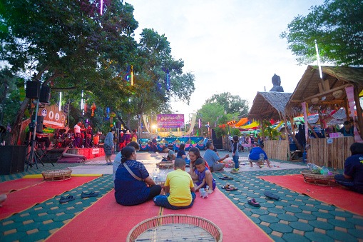 Early evening scene at traditional market in Ratchaburi. Market is seated next to temple  โบราณสถานวัดโขลง.  Market offers traditional thai food, thai clothes and other products and music and dance for public and visitors of market. People are sitting in center on the ground and are eating. In background is a stage and a banner for Loi Krathong beauty contest