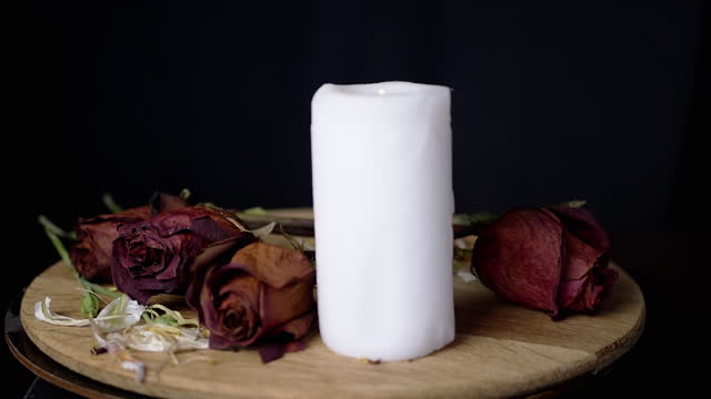 Hand Lights a White Candle against the Background of Rotating Dry Red Roses