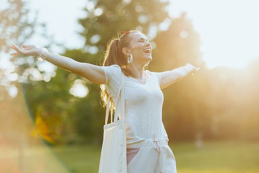 Summer time. smiling 40 years old woman in white shirt with tote bag rejoicing in the meadow outdoors.