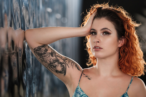 Tattooed young woman is leaning on a modern metallic wall. Cosmopolitan environment, the wall has a lot of reflections. Her hair is red and she has a piercing on her nose.