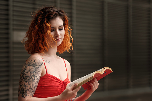 Caucasian model woman enjoying reading a book outdoors. She is smiling and she looks relaxed. Tattoos, piercing and red summer dress. Her hair is curly and red.