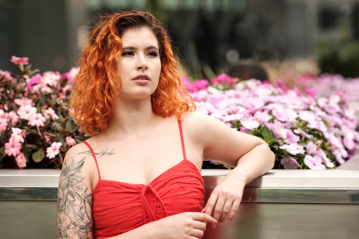 Attractive young woman leaning against flowers outdoors. She has a pensive look and she has a sensual mood. Urban environment, metallic place. She has tattoos and red hair.