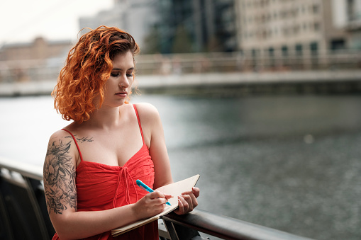 Caucasian woman writing on notebook and leaning on banister beside a canal in London. Inspiration and classic hobbies concept. No technology involved.