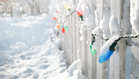 Idyllic snowy Xmas holiday background with wooden fence and snowed in sidewalk. Copy space. Selective focus on one light.