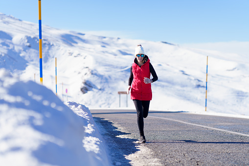 Full body of woman in outerwear and sunglasses jogging on asphalt road with snowy mountains