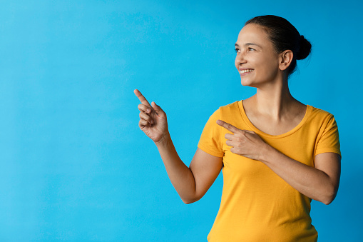 Caucasian female wearing a yellow t-shirt and is pointing  her fingers to the empty space on her left. She is standing in front of a blue wall.