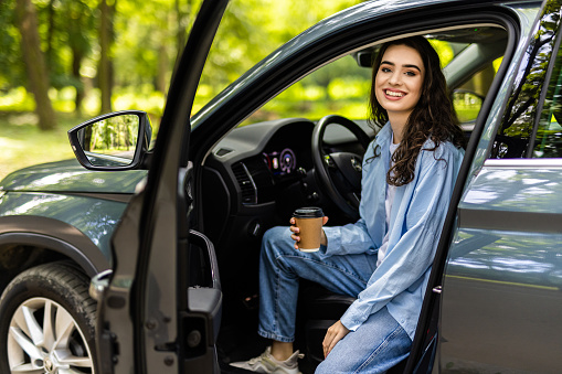 Beautiful businesswoman is sitting at steering wheel of her car. She is drinking coffee and smiling. The lady is looking at the camera happily