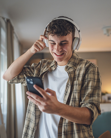 Portrait of teenage boy stand at home use headphones ans smartphone to play music or watch video podcast at home
