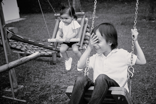 Black and white photography of brother and sister playing in swing in park. He's holding his hand on his head and smiling.