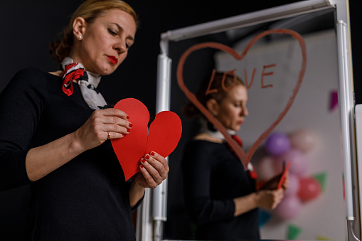 Low angle view of sad mid adult woman standing by the mirror with heart that she had drawn with a lipstick and tearing a paper heart in half down the middle.