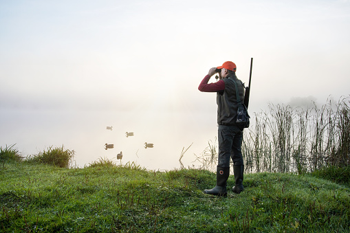 hunter with shotgun looking through binoculars near lake on foggy sunrise.