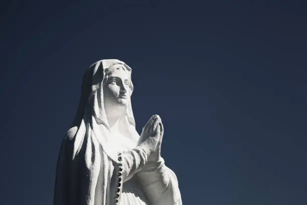 Photo of Ancient white statue of Virgin Mary against blue sky with free copy space for text.