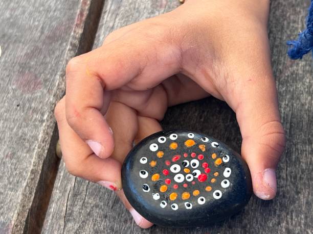 Horizontal high angle extreme closeup photo of a school student’s hand holding a small smooth colourful painted dot patterned stone on a rustic wood plank table. Armidale, New England high country, NSW.