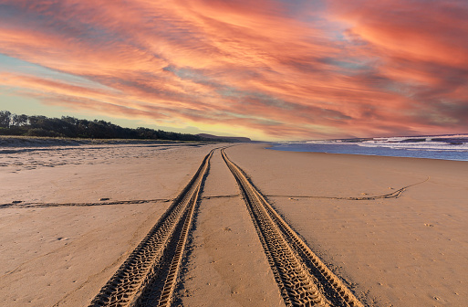 Landscape view of tire tracks on the sandy beach leading into the horizontal sea with orange sunlight ray effect on horizon view background.