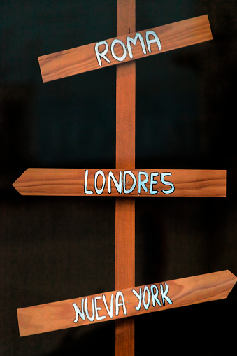 Wooden sign post, directional arrow symbol, black background. European capital cities in spanish language. Travel agency store window.