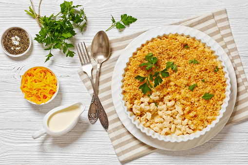 close-up baked mac and cheese sprinkled with panko breadcrumbs and fresh parsley in baking dish on white wooden table with ingredients, horizontal view, flat lay