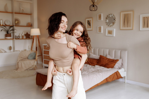 A young beautiful brunette mother plays with her little daughter, carries her on her back in the living room. The concept of unity, family and joint games with parents.