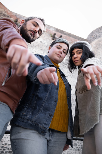 Two young women and a male friend, all with colored hair and piercings, look seriously at the camera, pointing their index fingers directly at the lens.