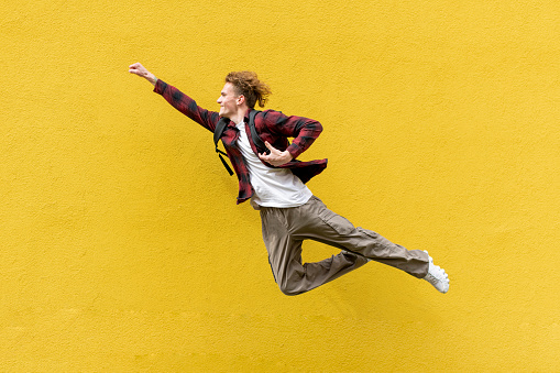 young guy student with backpack flies forward and hurries to study, man in superman pose jumps and runs in the air against yellow isolated wall, vacation and travel concept