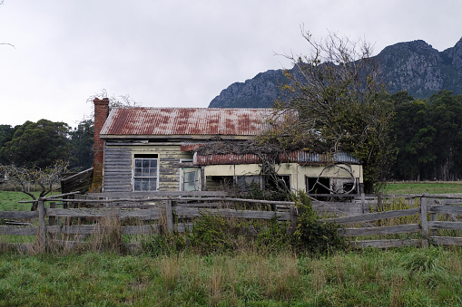 horizontal image of a quaint little old abandoned house with peeling paint and broken windows sitting behind a broken wooden fence under a beautiful blue sky with white clouds in the fall time.