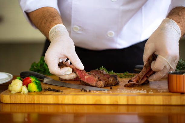 istock Chef cutting meat on a cutting board,Horizontal view of professional chef in black uniform salts raw steak on wooden chopped board. Backstage of preparing grilled pork meat at restaurant kitchen on dark blue background. Frozen motion. 1698373560
