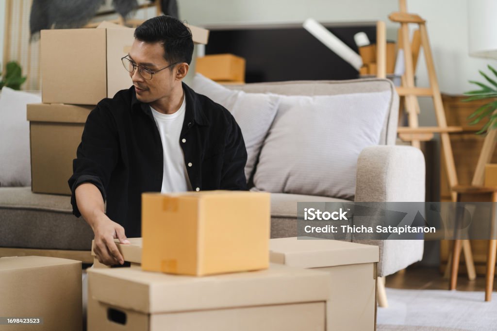 Tânăr care transportă cutii de carton ambalate cu obiecte personale în sufrageria confortabilă nemobilată în timpul zilei de mutare într-un apartament nou. - Fotografie de stoc Bărbat tânăr fără redevențe Tânăr care transportă cutii de carton ambalate cu obiecte personale în sufrageria confortabilă nemobilată în timpul zilei de mutare într-un apartament nou. - Fotografie de stoc Bărbat tânăr fără redevențe