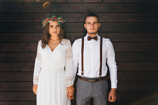 guy and girl standing near a wooden wall holding hands and looking at the camera. a girl in a white dress and a wreath on the head. guy with a beard in a bow tie and suspenders