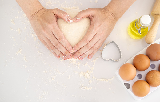 Cooking with love. Female hands holding dough in heart shape. Baking ingredients on the white table in home kitchen. Top view. Copy Space.