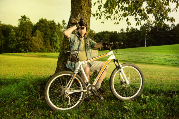 mid age cyclist is resting under tree and drink his last drops of water