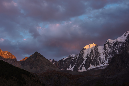 Atmospheric landscape with sunset gold reflection on huge snowy mountain top in violet dramatic sky. Giant snow mountains and pyramid shaped mountain in dusk. Snow-covered mountain range in twilight.