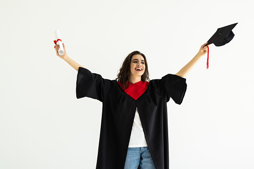 education, graduation and people concept - happy graduate student woman in mortarboard and bachelor gown with diploma celebrating and making winning gesture over white background