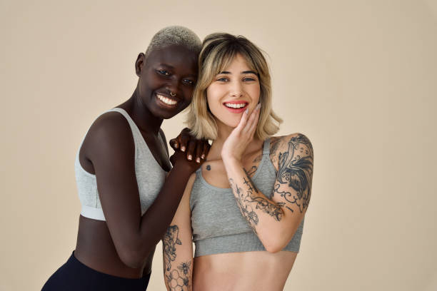 Two happy diverse fit women wearing sport tops standing on background, portrait. Two happy diverse fit women, Black and European young sporty gen z girls friends multiethnic models wearing sportswear tops standing smiling looking at camera on beige background, portrait. black tattoo stock pictures, royalty-free photos & images
