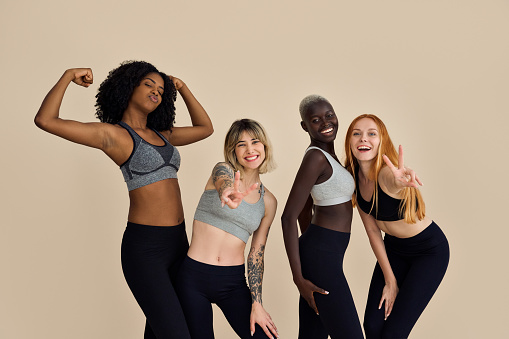 Happy fit sporty diverse different girls group having fun posing at beige background. Multiracial positive young women friends wear sportswear advertising fitness gym yoga body trainings together.