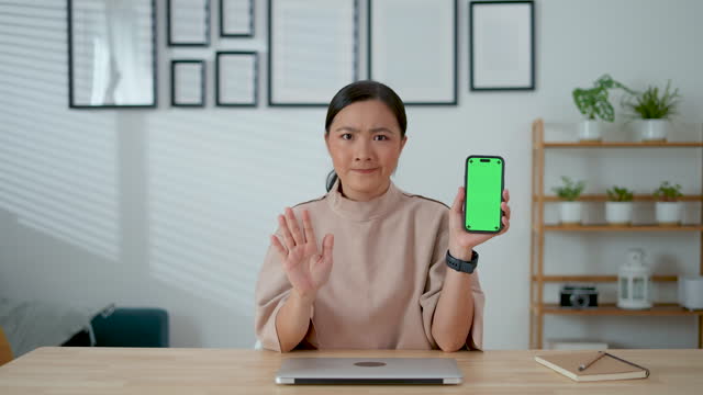 Asian woman feel upset showing stop sign by hand and holding smart phone showing green screen vertical orientation, sitting at home office.