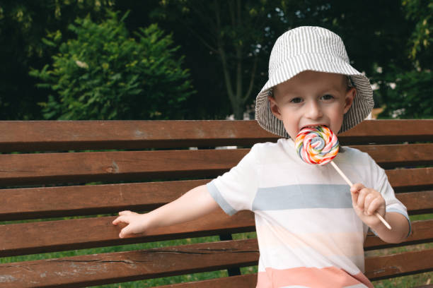 istock A boy in panama gnaws a multi-colored lollipop on a bench. Sweets are bad for teeth. 1698775013