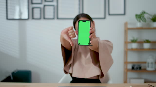 Asian woman feel upset showing thumb down and holding smart phone showing green screen vertical orientation, sitting at home office.