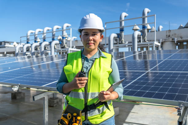 Female Asian engineer with protective workwear working outdoor holding communication device and digital tablet including scientific instrument for inspecting solar cell panels, smiling engineer supporting employee engagement